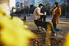 Man and woman holding drinks smiling and having a conversation. Man is sitting down on bench.