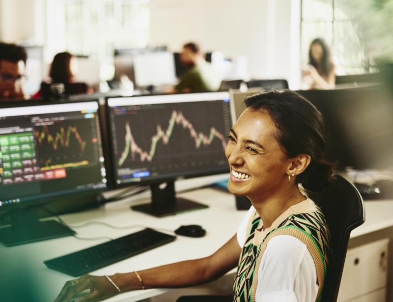 Woman smiling while sitting behind her desk, screens displaying financial information.