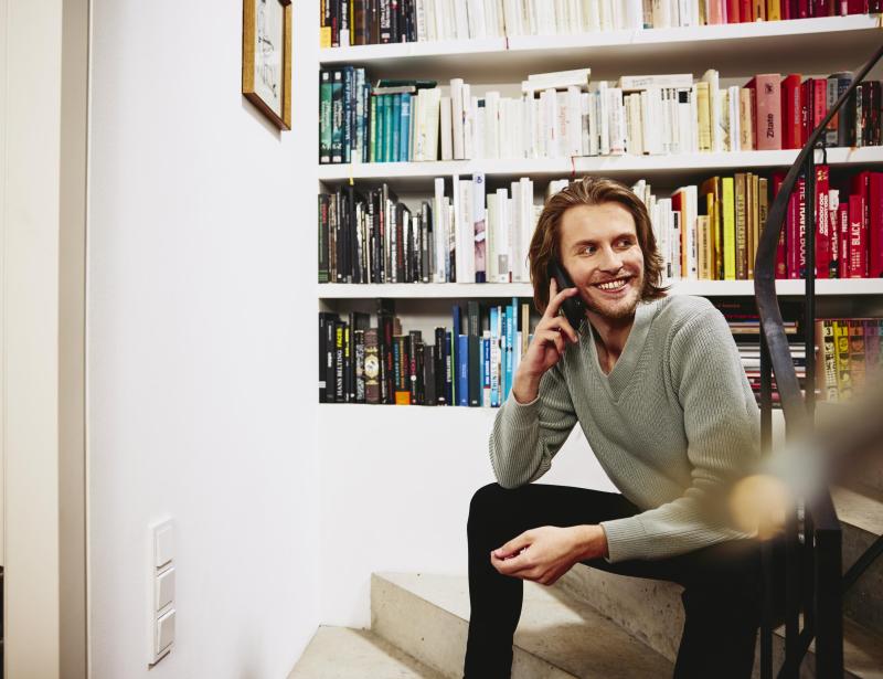 Smiling man sitting on stairs, having a phonecall. bookshelf in background