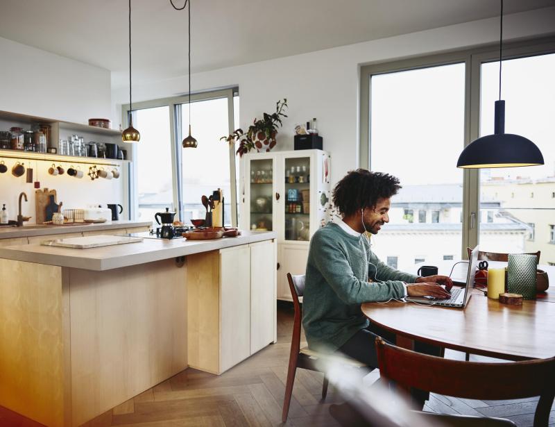 Smiling man sitting at his dining table working on his laptop. Kitchen in the background.