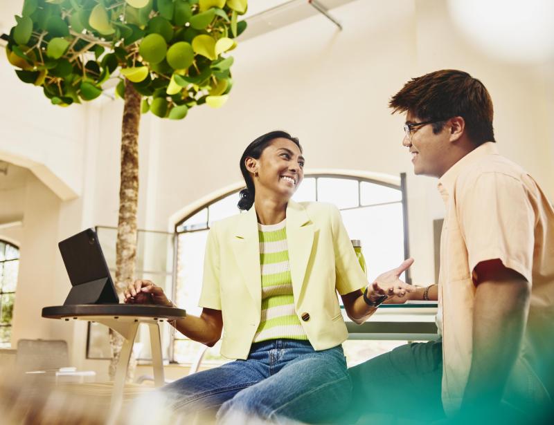 Man and woman sitting at table having a chat, while woman working on a tablet.