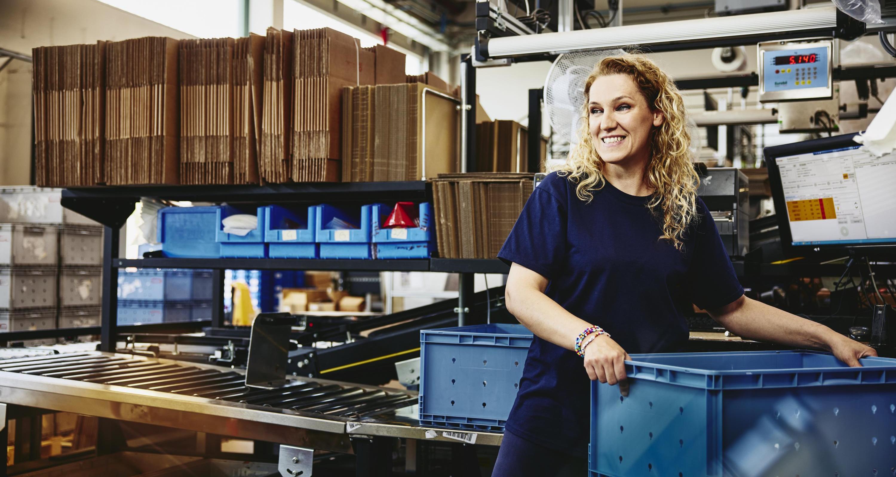 Smiling woman standing next to conveyer belt holding a crate